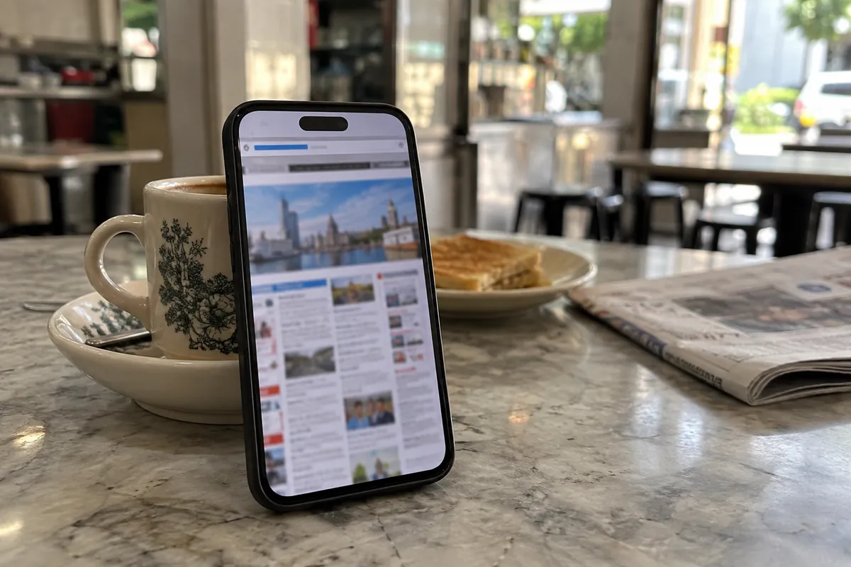 Phone propped against a kopitiam coffee cup showing a cramped desktop-style website that doesn't fit the mobile screen